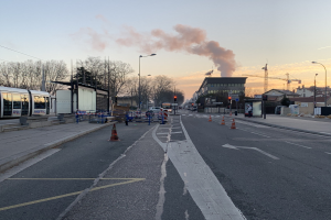 Eine Stadtstraße mit Absperrbaken, einem Bus, Laternenpfählen, Bäumen, Gebäuden, Kränen und Rauch, der aus einem entfernten Gebäude aufsteigt.