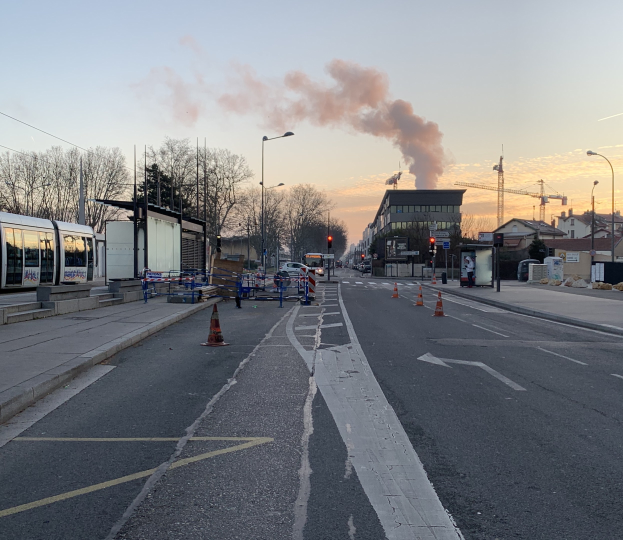 Eine Stadtstraße mit Absperrbaken, einem Bus, Laternenpfählen, Bäumen, Gebäuden, Kränen und Rauch, der aus einem entfernten Gebäude aufsteigt.