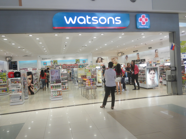 Eine Watsons Apotheke in einem Einkaufszentrum, mit Menschen auf dem Boden, Gegenständen auf Regalen, Schildern mit Text, einer Wanduhr und Deckenleuchten.
