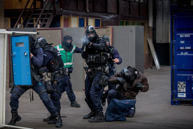 Polizeibeamte in Einsatzausrüstung stehen vor einem Gebäude in einer Reihe, einige halten Waffen, mit einer Person im Vordergrund und einem blauen Objekt mit Text auf der rechten Seite.