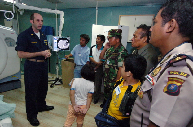Eine Gruppe von Menschen in einem Krankenhauszimmer mit einem Mann, der ein Mikrofon hält, einem Bett mit einer Decke, medizinischen Geräten, einem Röntgenständer, einer Texttafel, einer Tür und Deckenleuchten.