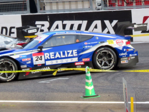 Ein blau-weißer Rennwagen fährt auf einer Rennstrecke, mit einem Verkehrskegel im Vordergrund und Schildern, Geländern und anderen Gegenständen im Hintergrund.