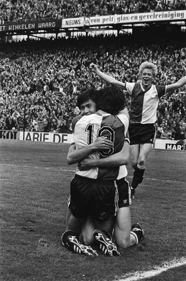 Drei Fussballspieler umarmen sich auf dem Boden beim Feiern eines Tors, mit jubelnder Menge und schwenkenden Fahnen im Hintergrund, dargestellt in Schwarz-Weiß.