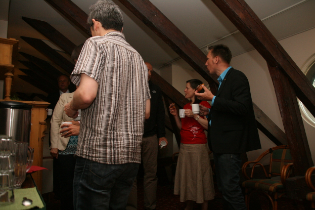 Eine Gruppe von Menschen mit Tassen in der Hand, links ein Becher und ein Tisch, oben ein Dach und rechts Stühle.