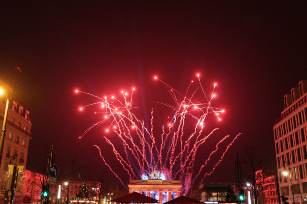 Eine Stadtstraße in Berlin an Silvester, gefüllt mit Gebäuden, Bäumen, Laternen, Verkehrszeichen, Zeltplanen und Menschen, mit einem Feuerwerk erleuchteten Himmel im Hintergrund.