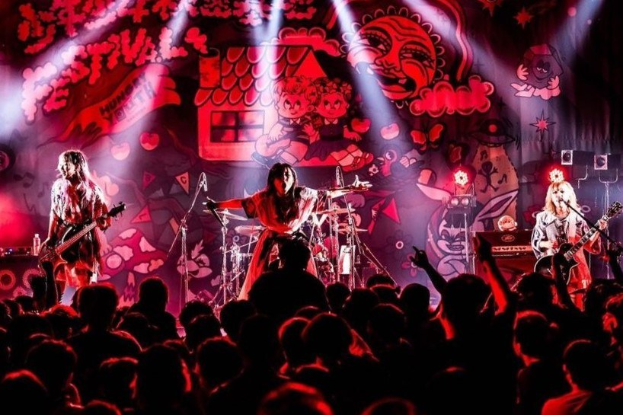 Ein Live-Auftritt der japanischen Rockband Korn auf der Bühne mit Musikern, die Gitarren spielen und in Mikrofone singen, ein beleuchtetes Gemälde an der Wand dahinter und eine sichtbare Menge unten.