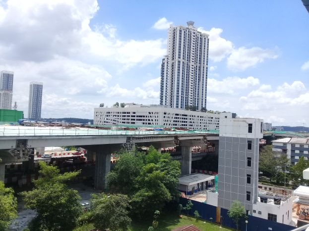 Luftaufnahme einer Stadt mit Gebäuden, Bäumen, Gras, Fahrzeugen, einer Brücke, Pfählen und einem bewölkten Himmel, die eine Baustelle für einen neuen Wohnkomplex zeigt.