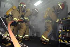 Feuerwehrleute in Schutzausrüstung mit Helmen und Sauerstoffflaschen arbeiten an einem Löschfahrzeug, mit einem Gestell voller Gegenstände und Hintergrundrohren und Lichtern.