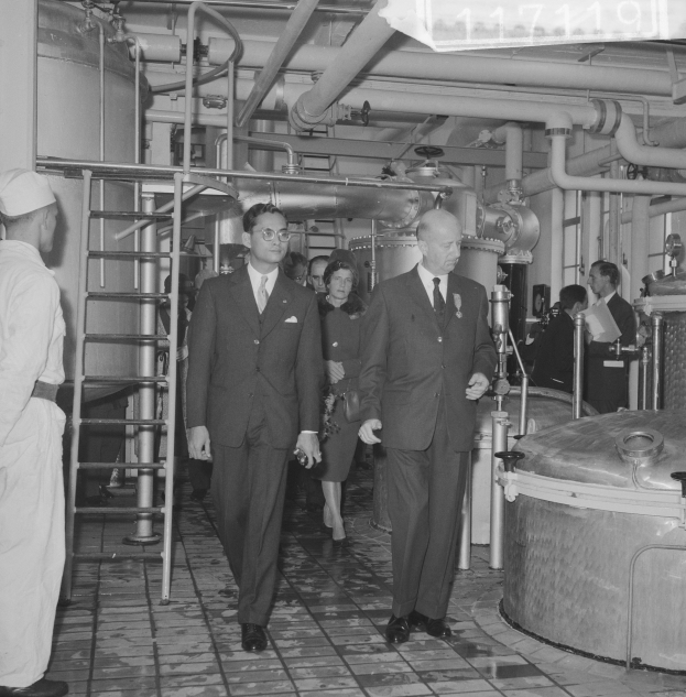 Eine Gruppe von Männern in Anzügen und Krawatten durch eine schwarz-weiße Fabrik mit einer Leiter auf der linken Seite und Maschinen im Hintergrund.
