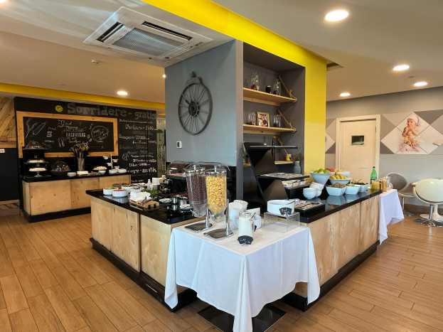 Ein Restaurant-Innenraum mit Tischen und Stühlen, Essensbehältern, einer Tafel mit Text, einer Wanduhr, Deckenleuchten, einer Klimaanlage und einer Tür im Hintergrund, gelegen im Hôtel de Ville in Paris.