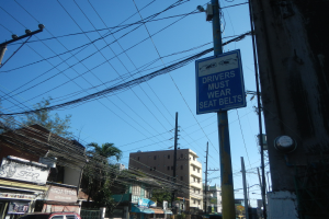 Stadtstraße mit fahrenden Autos, Strommäste mit Drähten, Gebäude, Bäume und Schilder; ein Schild an einem Strommast lautet "Fahrer müssen Sitzgürtel tragen".