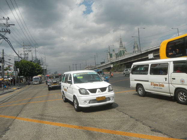 Eine belebte Stadtstraße mit Autos, Bussen, einem Minibus, Fußgängern auf dem Gehweg, Strommasten, Laternen, Bäumen und einer Brücke im Hintergrund bei bewölktem Himmel.