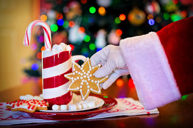 Mann in einem Santa Claus Hut, der einen Becher heiße Schokolade und einen Bonbonstab hält und ein Teller mit Plätzchen und ein Tuch auf dem Tisch vor ihm hat, vor einem Weihnachtsbaum und einer Wand.
