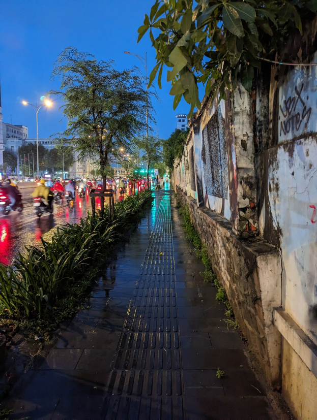 Eine nächtliche Stadtstraße mit Graffiti an der Seite, beleuchtet von Straßenlaternen, mit fahrenden Kraftfahrzeugen, Bäumen und Pflanzen an den Seiten, Gebäuden im Hintergrund und einem sternenklaren Himmel.