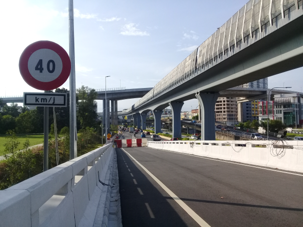 Eine Autobahn mit einem Geschwindigkeitsbegrenzungsschild, Fahrzeuge, eine Brücke mit Pfeilern, Laternen, Bäume, Gebäude und ein bewölkter Himmel.