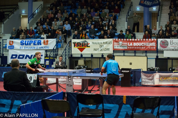 Zwei Männer spielen Tischtennis in einem Innenraum, umgeben von Zuschauern, leeren Stühlen und Werbetafeln, mit Text in der linken unteren Ecke.