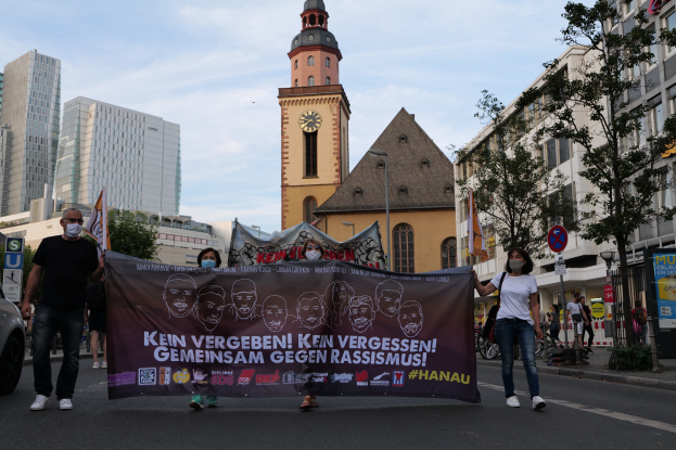 Eine Gruppe von Menschen mit Masken, die eine Plakette tragen, geht eine Straße entlang, mit einem geparkten Auto auf der linken Seite, Gebäude und einen Kirchturm im Hintergrund unter einem klaren blauen Himmel.