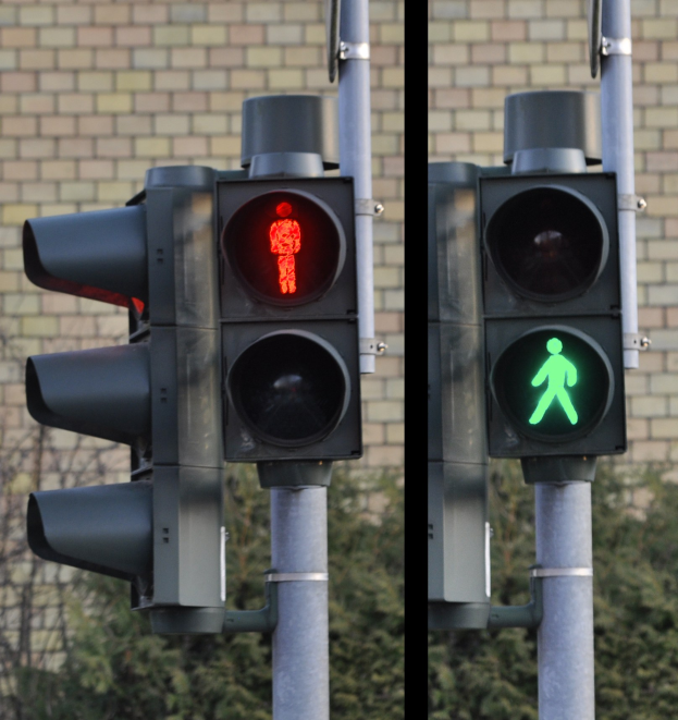 Zwei Ampeln an einem Mast mit roter und grüner Lampe vor einer Backsteinwand mit Bäumen im Hintergrund.