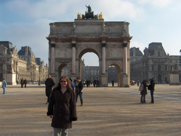 Eine Frau in einem Mantel lächelt, während sie auf einer Straße steht, mit Menschen im Hintergrund, die stehen und gehen, sowie Statuen auf der Architektur und Gebäude unter einem blauen Himmel mit Wolken.