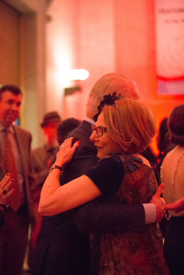 Zwei Menschen umarmen sich auf einer Party, während andere in der Nähe stehen und eine beleuchtete Wand im Hintergrund zu sehen ist.