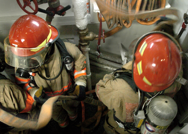 Feuerwehrleute in Helmen und Schutzausrüstung arbeiten an einem Feuerhydranten mit Rohren, Ventilen und anderen Gegenständen im Hintergrund, mit Text unten im Bild.
