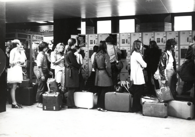 Eine Gruppe von Menschen mit Gepäck, einige halten Taschen, steht in einer schwarz-weißen Flughafen-Szene mit Schließfächern im Hintergrund.