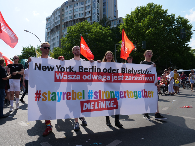 Eine Gruppe von Menschen marschiert auf einer von Bäumen gesäumten Straße in Berlin, Deutschland, mit einer Tafel, auf der "Stay Rebel Stronger Together" steht, während Gebäude und Laternenpfähle zu sehen sind und ein bewölkter Himmel über ihnen liegt.