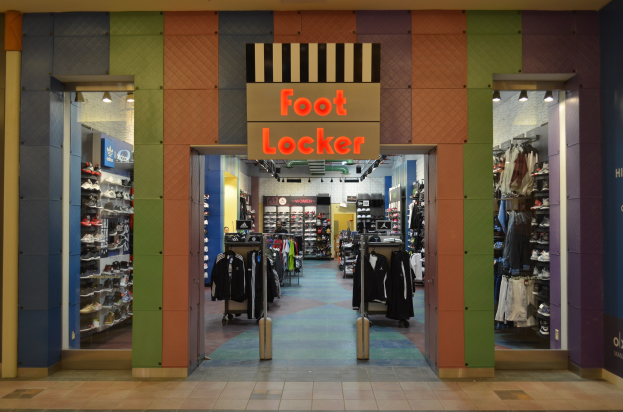 Innenansicht eines Foot Locker-Schuhgeschäfts mit Kleidung an Bügeln und Ständern unter Deckenbeleuchtung, wobei der Geschäftsboden sichtbar ist.
