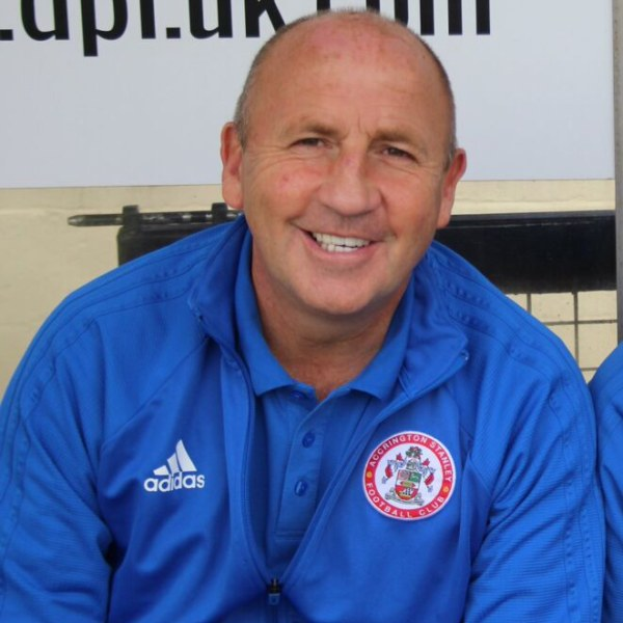 Zwei Männer in einem Raum, einer in einer blauen Jacke neben dem anderen sitzend, beide lächeln; eine Tafel ist an der Wand hinter ihnen angebracht.