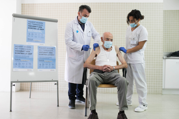 Alter Mann erhält eine Impfung von zwei maskierten Ärzten in einem Krankenhauszimmer, mit einer Tafel und medizinischem Zubehör in der Nähe.