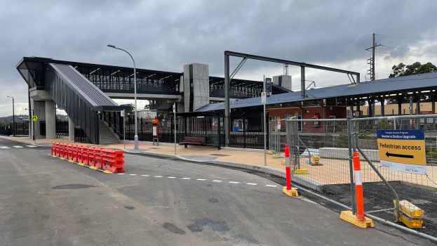 Ein Bahnhofsbild mit einer Brücke im Hintergrund, Straßenlaternen, Strommasten, Stromkabel, Bäume, Gebäude, Fahrzeuge auf der Straße, Absperrpoller, Schilder, Bänke und ein bewölkter Himmel.