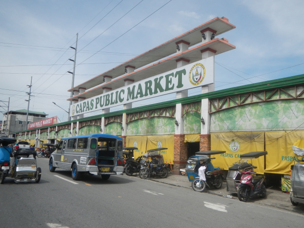 Eine belebte Stadtstraße mit Autos, Motorrädern und Rikschas, die an einem Gebäude mit der Aufschrift "Capas Public Market" vorbeifahren, vor einem Hintergrund aus Strommasten, Gebäuden und bewölktem Himmel.