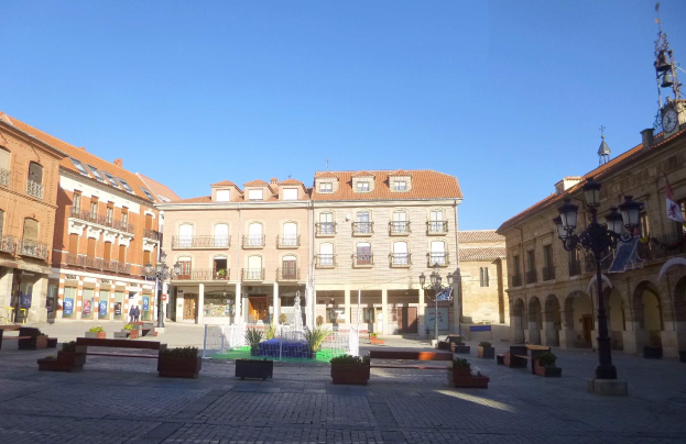 Ein Rathausplatz in einer Stadtmitte mit einem zentralen Brunnen, umgeben von Bänken, Topfpflanzen, Straßenlaternen, Straßenlaternen, einem Uhrenturm und Gebäuden mit Fenstern unter einem klaren blauen Himmel.
