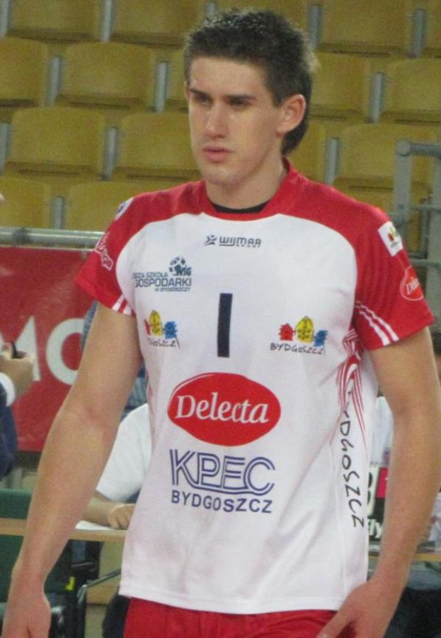 Ein Mann in rot-weißer Handballdressur steht auf einem Feld, entschlossen schauend, mit Zuschauern und einem Banner mit 'Krasnoyarsk vs Bydgoszcz' im Hintergrund.