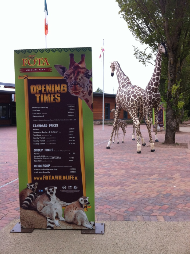 Giraffen stehen neben Bäumen, Schuppen und Fahnen, mit einer Tafel, die Text, Logos und Tierbilder anzeigt.