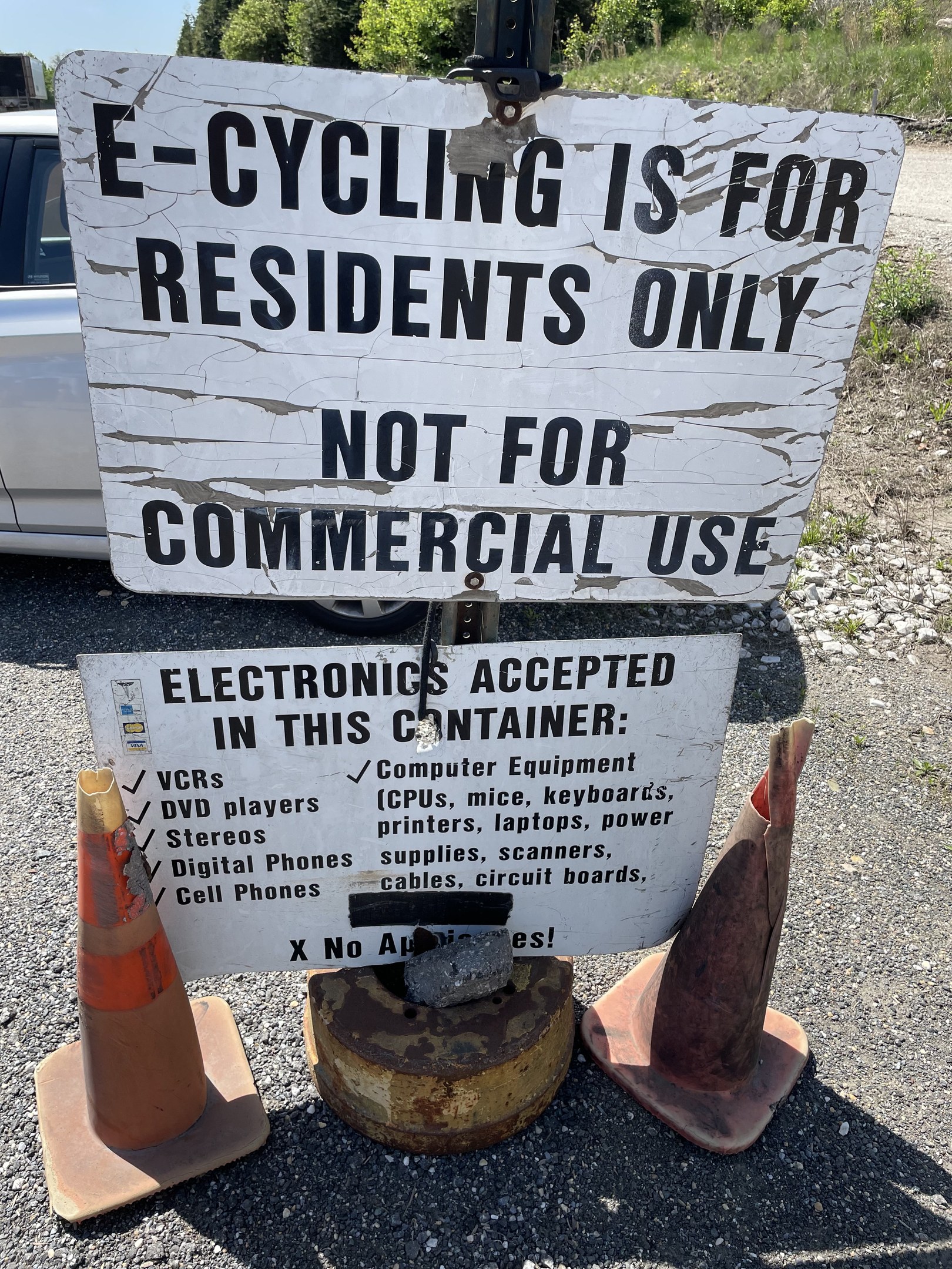 Schild mit der Aufschrift "e-Bike-Fahren nur für Anwohner" neben einer Straße, umgeben von Absperrgittern und anderen Gegenständen, mit einem Fahrzeug, Bäumen und einem klaren blauen Himmel im Hintergrund.