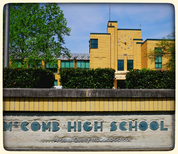 Eine Wand mit "MCCOMB HIGH SCHOOL" darauf, mit Pflanzen, Bäumen, einem Pfahl, einem Gebäude und einem bewölkten Himmel im Hintergrund.