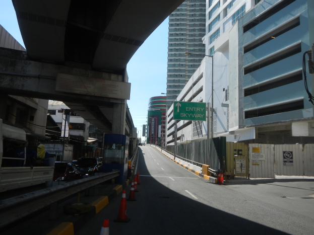 Stadtstraße mit Verkehrskegeln am Straßenrand, Fahrzeuge auf der Straße, eine Brücke links, Gebäude mit Namensschildern auf beiden Seiten, Laternenpfähle und ein Himmel im Hintergrund.