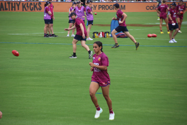 Frauen spielen Australian Rules Football auf einem Rasenfeld, mit verstreuten Bällen und einem Banner im Hintergrund, einige tragen Kappen und Turnschuhe.