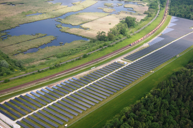 Luftaufnahme eines Solarparks mit Panels, einem Zug auf einer Schiene, umgeben von Bäumen, Gras, Pflanzen und Wasser.