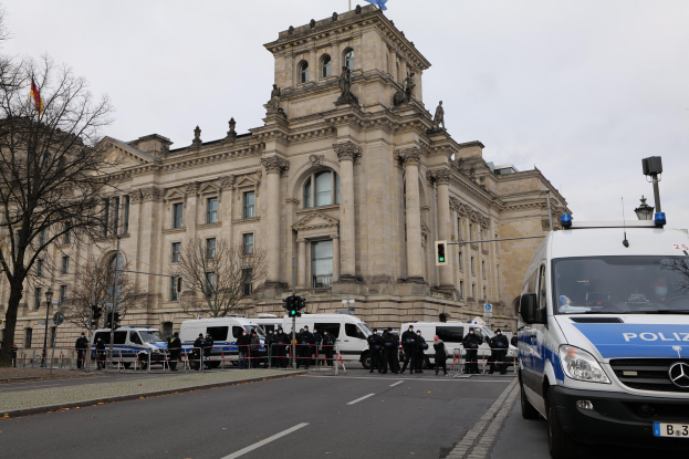 Eine Gruppe von Polizisten steht vor dem Reichstaggebäude in Berlin, Deutschland, mit Fahrzeugen, einem Zaun, Verkehrsampeln, Laternenmasten, Bäumen und Fahnen im Hintergrund, unter einem klaren Himmel.