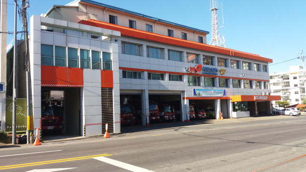 Feuerwache mit einem Feuerwehrauto davor, umgeben von Gebäuden, Fahrzeugen, Verkehrskegeln, Strommasten, Kabeln, Schildern, Bäumen und einem klaren Himmel.