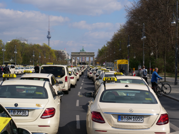 Eine lange Reihe von Taxis, die an einer belebten Straße in Berlin, Deutschland, geparkt sind, mit Radfahrern und Fußgängern auf dem Gehweg, flankiert von Laternen und Bäumen, und Gebäuden, einem Bogen und einem Turm im Hintergrund bei einem bewölkten Himmel.