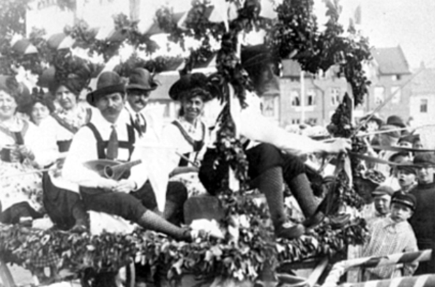 Eine Gruppe von Menschen in traditioneller bayrischer Tracht fährt auf einem mit Blättern geschmückten Festwagen in einem Umzug, einige halten Musikinstrumente, mit Gebäuden und einem klaren Himmel im Hintergrund, dargestellt in Schwarz-Weiß.