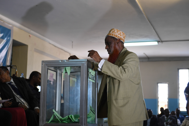 Ein Mann steht vor einer Glasurne mit Papieren, umgeben von sitzenden Wählern, mit einem Banner an der linken Wand und Fenstern im Hintergrund.