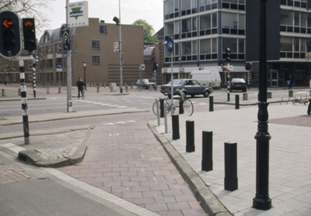 Eine Stadtstraßenszene mit einem geparkten Fahrrad, fahrenden Fahrzeugen, Fußgängern, Pfosten, Verkehrsampeln, Verkehrszeichen, Bäumen, mehrstöckigen Gebäuden und einem klaren blauen Himmel.