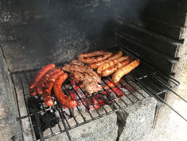 Grill mit Würstchen und Fleisch darauf, umgeben von brennender Kohle und Flammen, auf Gras im Freien