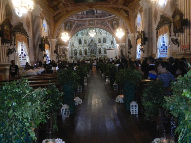 Eine Hochzeitszeremonie in einer Kirche mit Gästen auf den Bänken und Stehplätzen, Topfpflanzen im Gang, Skulpturen an den Wänden, Vorhänge an den Fenstern, Kronleuchter von der Decke und ein Bildschirm im Hintergrund.