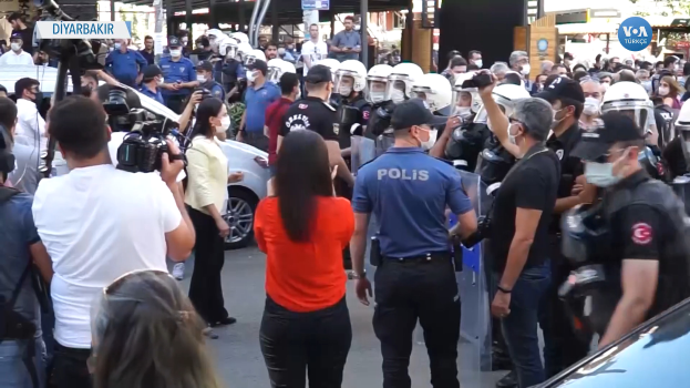 Eine Gruppe von Menschen steht vor einer Reihe von Polizisten in Helmen und mit Kameras, mit Fahrzeugen auf der Straße und Gebäuden, Bäumen und Schildern im Hintergrund, die "türkische Polizei setzt sich gegen Demonstranten in Ankara ein" zeigen.
