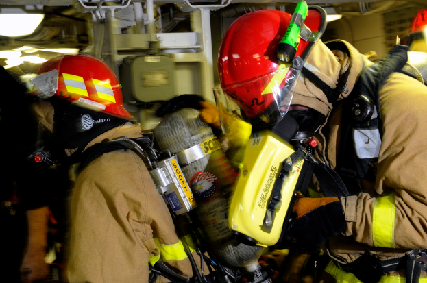 Eine Gruppe von Feuerwehrleuten in voller Ausrüstung, einschließlich Helmen und Sauerstoffflaschen, arbeitet an einem Flugzeug während eines Übungsmanövers, mit verschiedenen Gegenständen und Lichtern im Hintergrund.